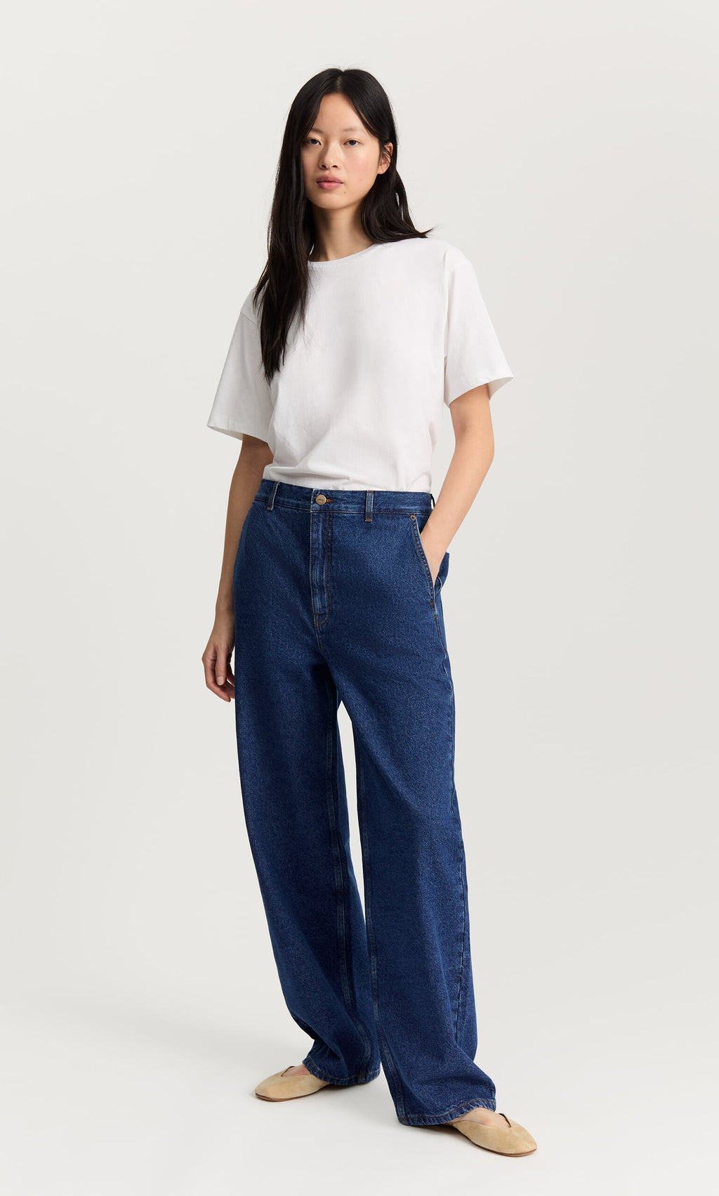 Plümo- Woman wearing Carlo Jeans and a white tee with beige flats stands against a plain white background.