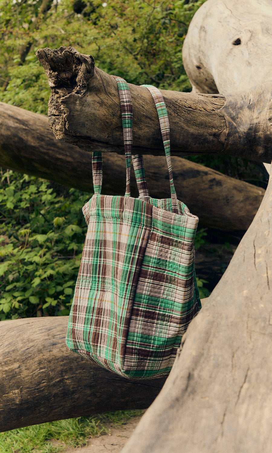 Plümo- The Checked Quilted Bag in green, brown, and white hangs on a large fallen tree trunk outdoors.