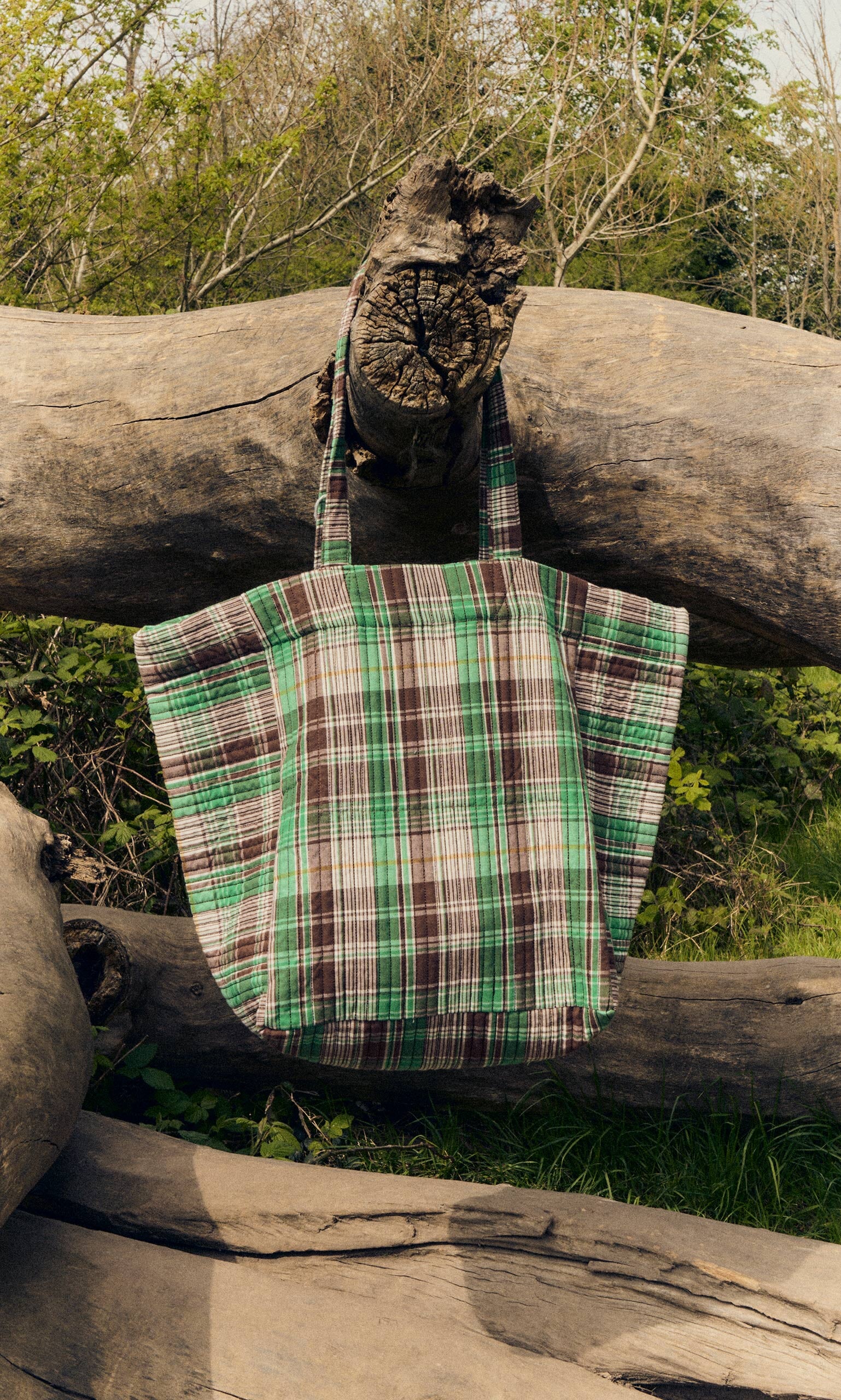 Plümo- The Checked Quilted Bag in green, brown, and white checks hangs on a large fallen tree outdoors.