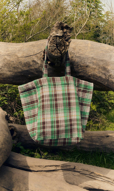 Plümo- The Checked Quilted Bag in green, brown, and white checks hangs on a large fallen tree outdoors.