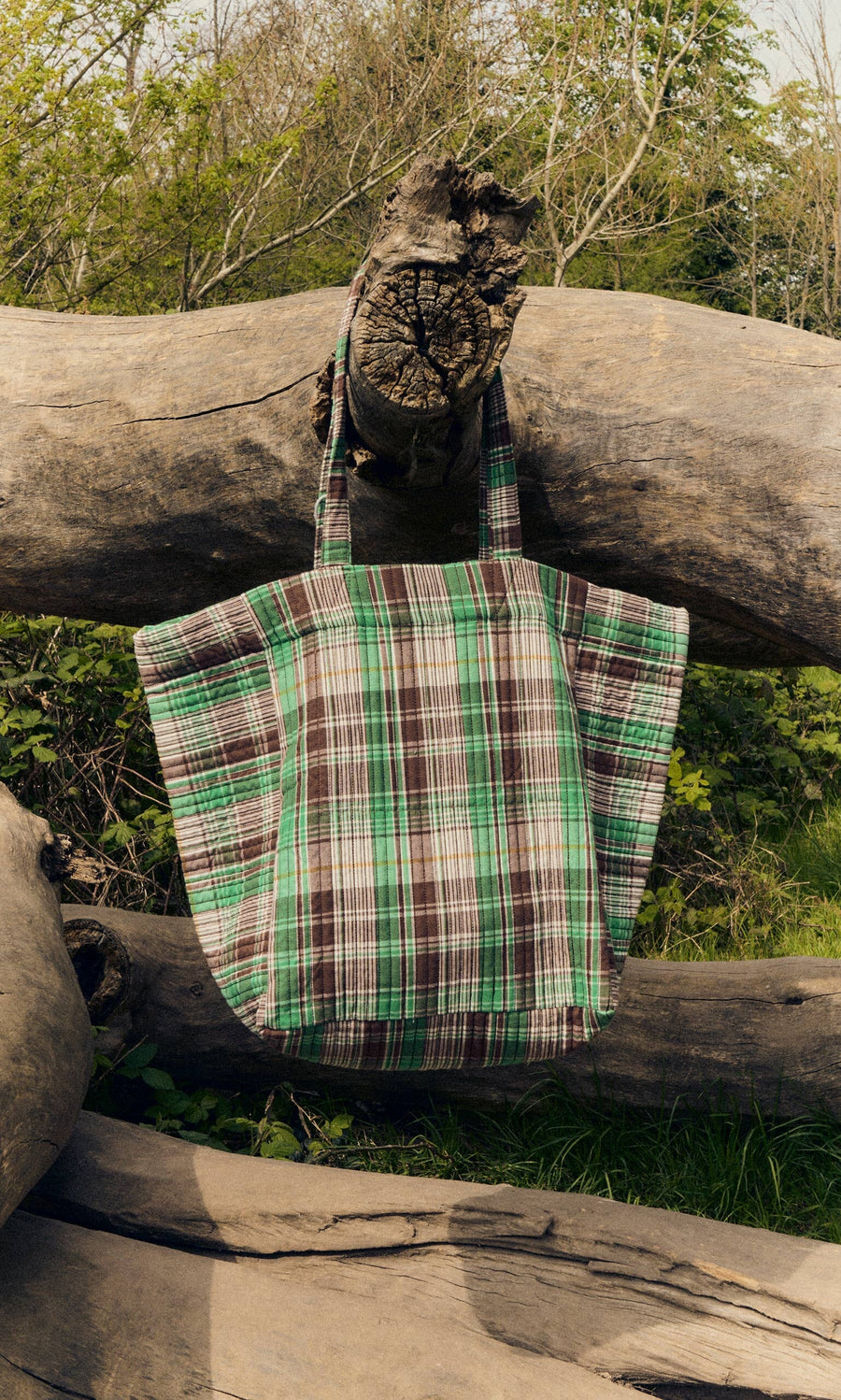 Plümo- The Checked Quilted Bag in green, brown, and white checks hangs on a large fallen tree outdoors.