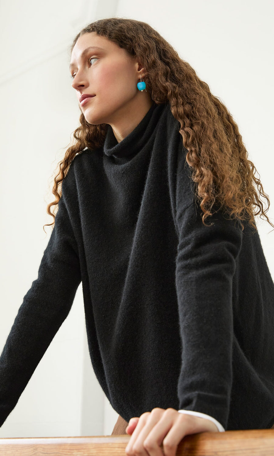 Plümo- Woman with long curly hair wears the Onyx Rollneck, leaning forward in this classic wardrobe staple.