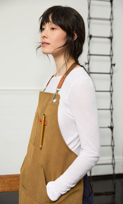 Plümo- A woman in a brown apron holds the Workshop Pen, standing sideways with her hand in her pocket, looking ahead.