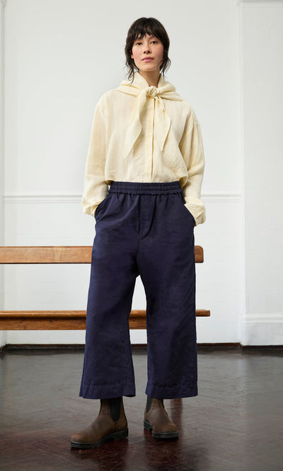 Plümo- Woman indoors in a cream blouse, Cromer Pants - Navy, and brown boots.