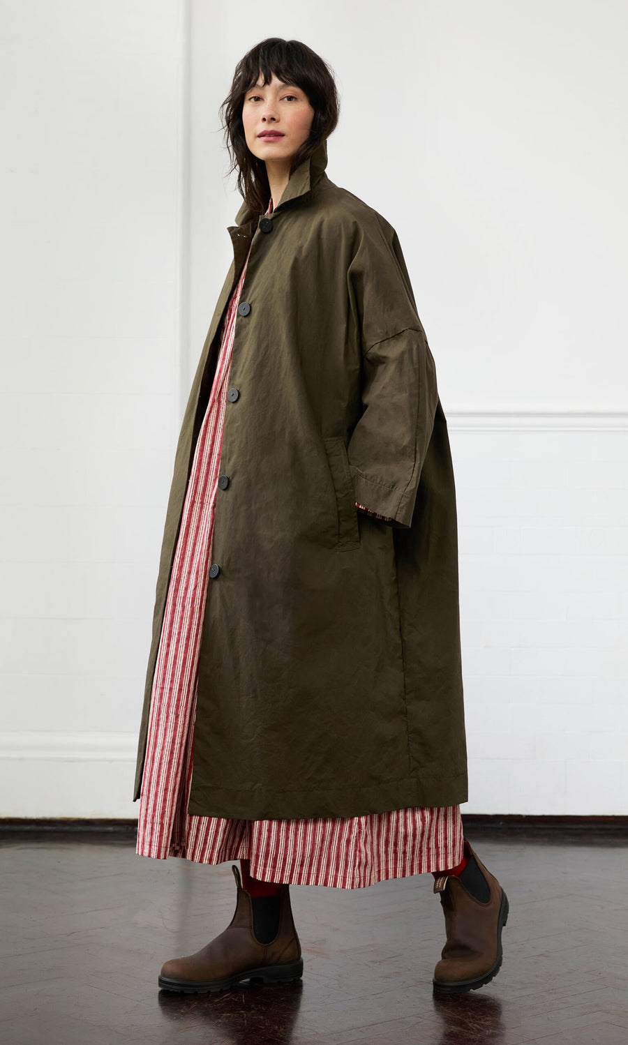 Plümo- A woman in a Doherty Coat - Olive, inspired by vintage workwear, over a red and white striped dress stands indoors.