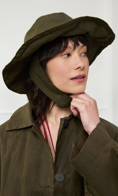 Plümo- Woman in a Gaucho Waxed Hat and matching olive coat looks to the side, touching her chin with one hand.
