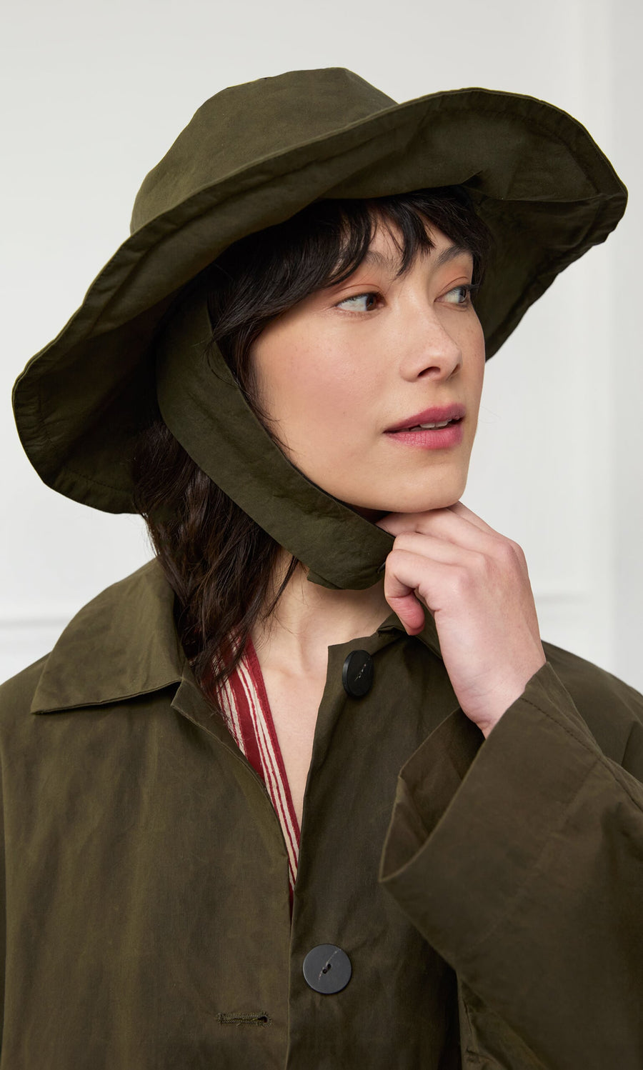 Plümo- Woman in a Gaucho Waxed Hat and matching olive coat looks to the side, touching her chin with one hand.