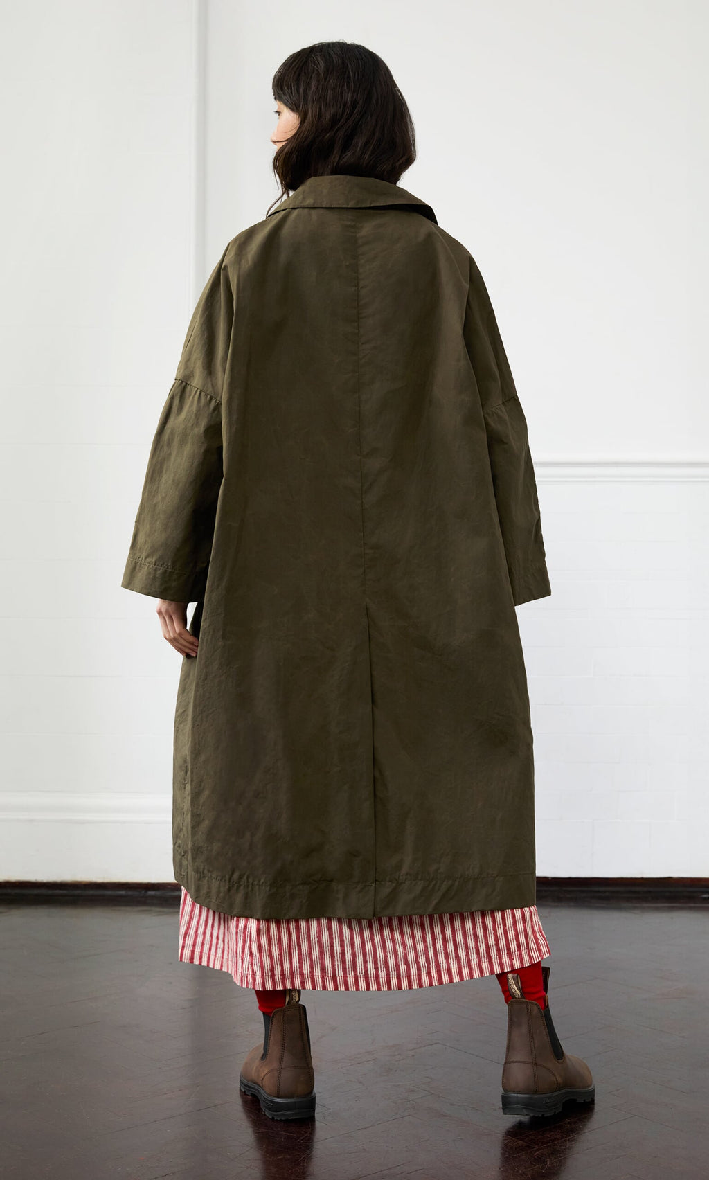Plümo- Person in the Doherty Coat - Olive, red and white striped skirt, and brown boots stands on a dark floor, facing away.