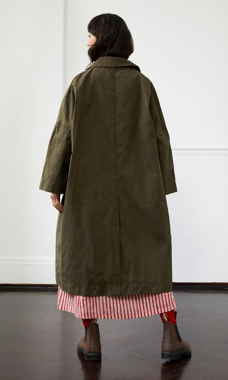 Plümo- Person in the Doherty Coat - Olive, red and white striped skirt, and brown boots stands on a dark floor, facing away.