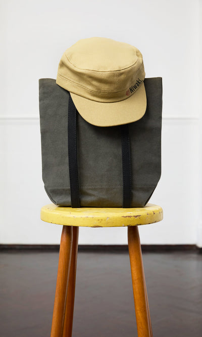 Plümo- A Niwaki Cap with an adjustable fit sits on a gray tote bag atop a yellow wooden stool.
