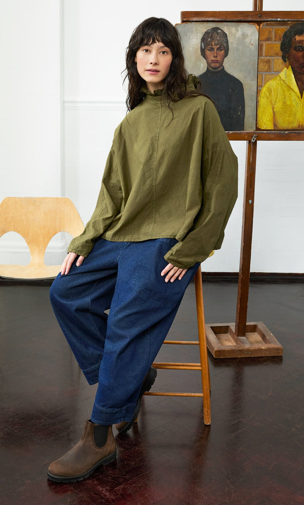 Plümo- Someone in a green jacket and the Frieda Blouse with a ruffled neck sits on a stool in an art studio with paintings behind.