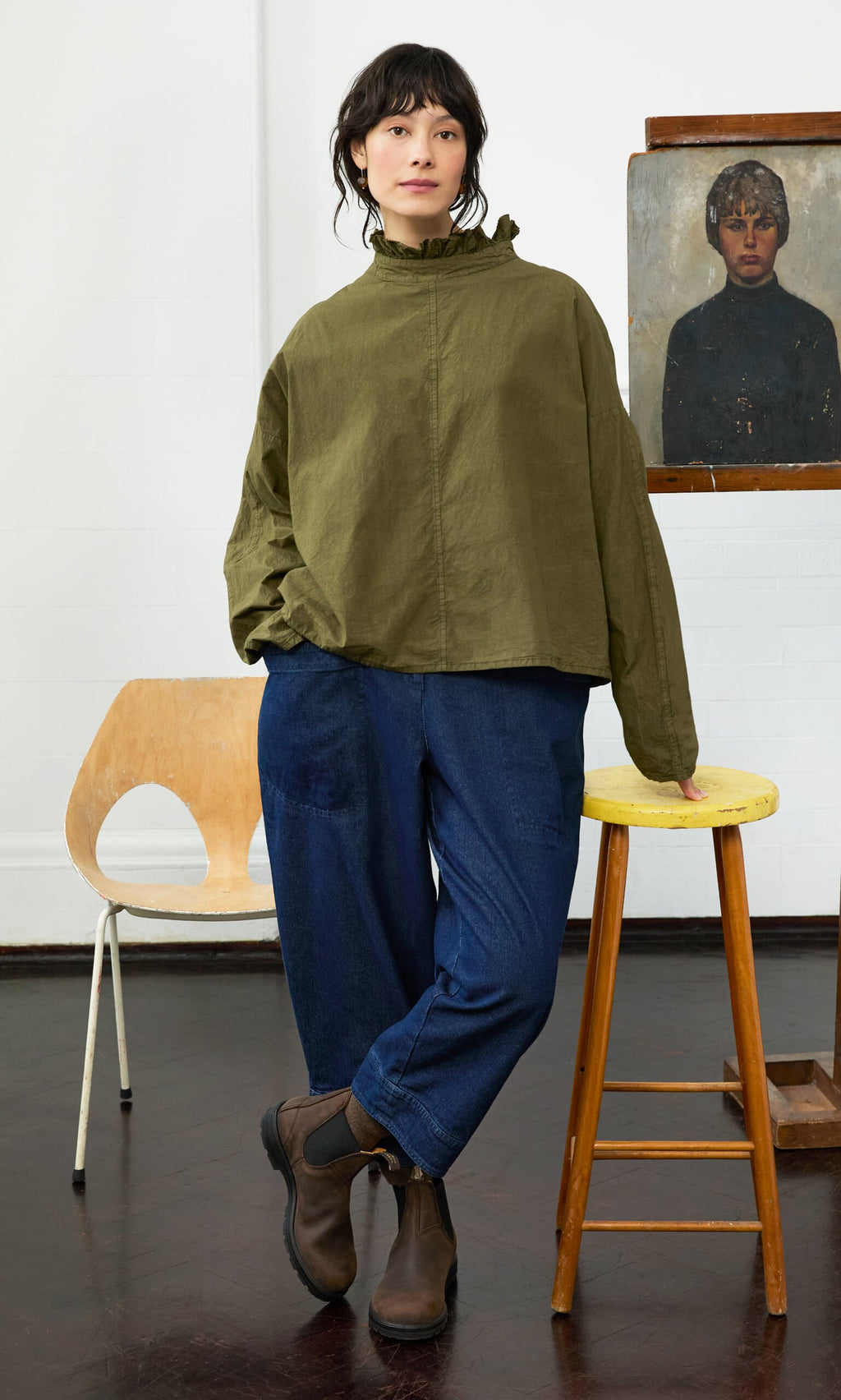 Plümo- Woman in the Frieda Blouse and blue pants leans on a yellow stool near a chair and portrait painting.
