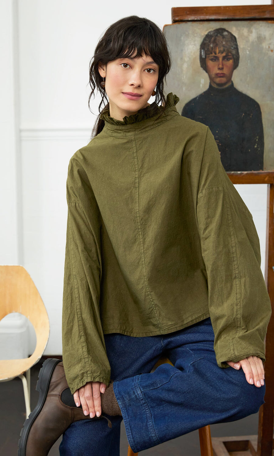 Plümo- Woman wears the Frieda Blouse with blue jeans indoors, posing in front of a painting of a woman.
