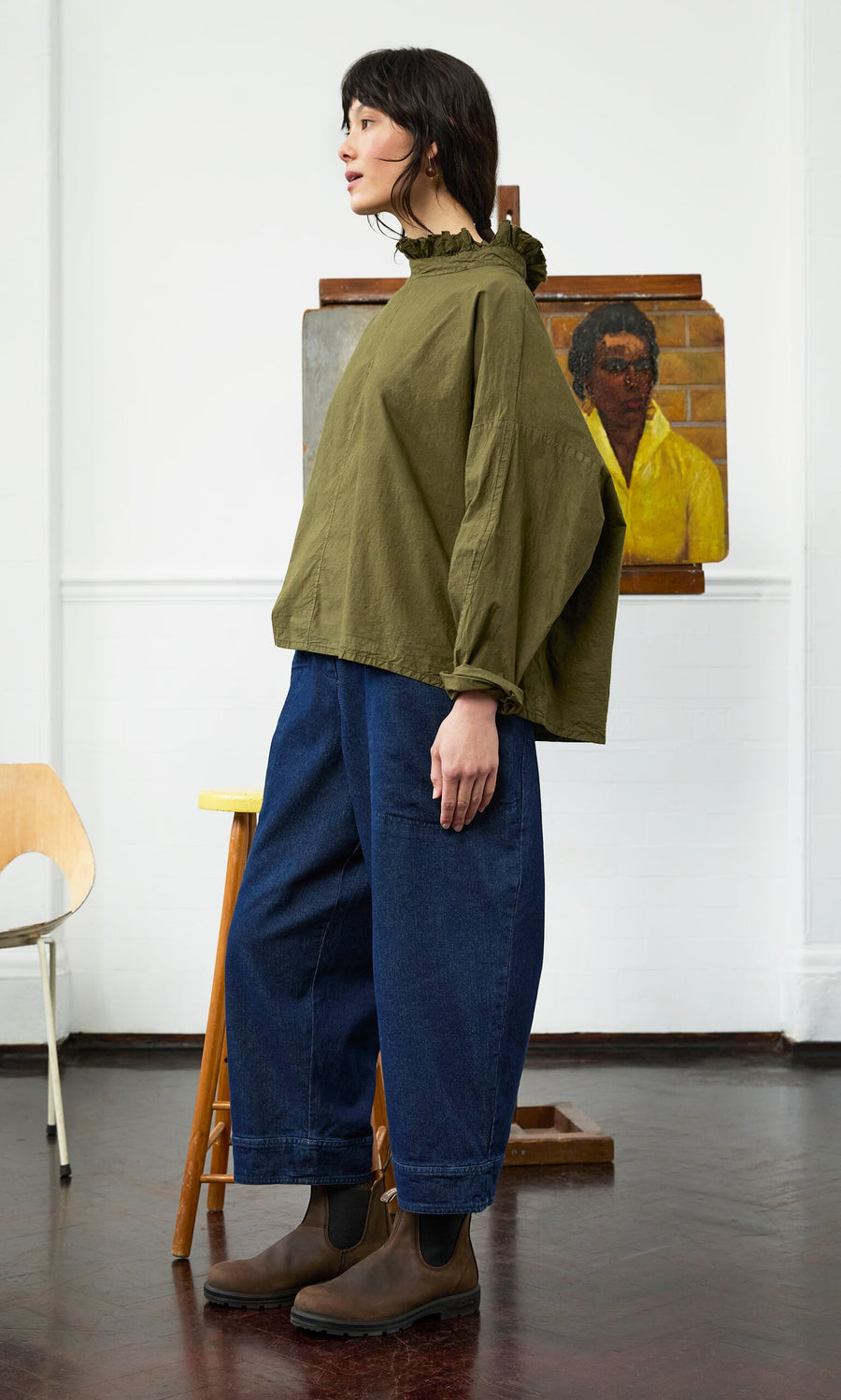 Plümo- A woman in a relaxed Frieda Blouse and blue jeans stands sideways in an art studio with a portrait painting behind her.