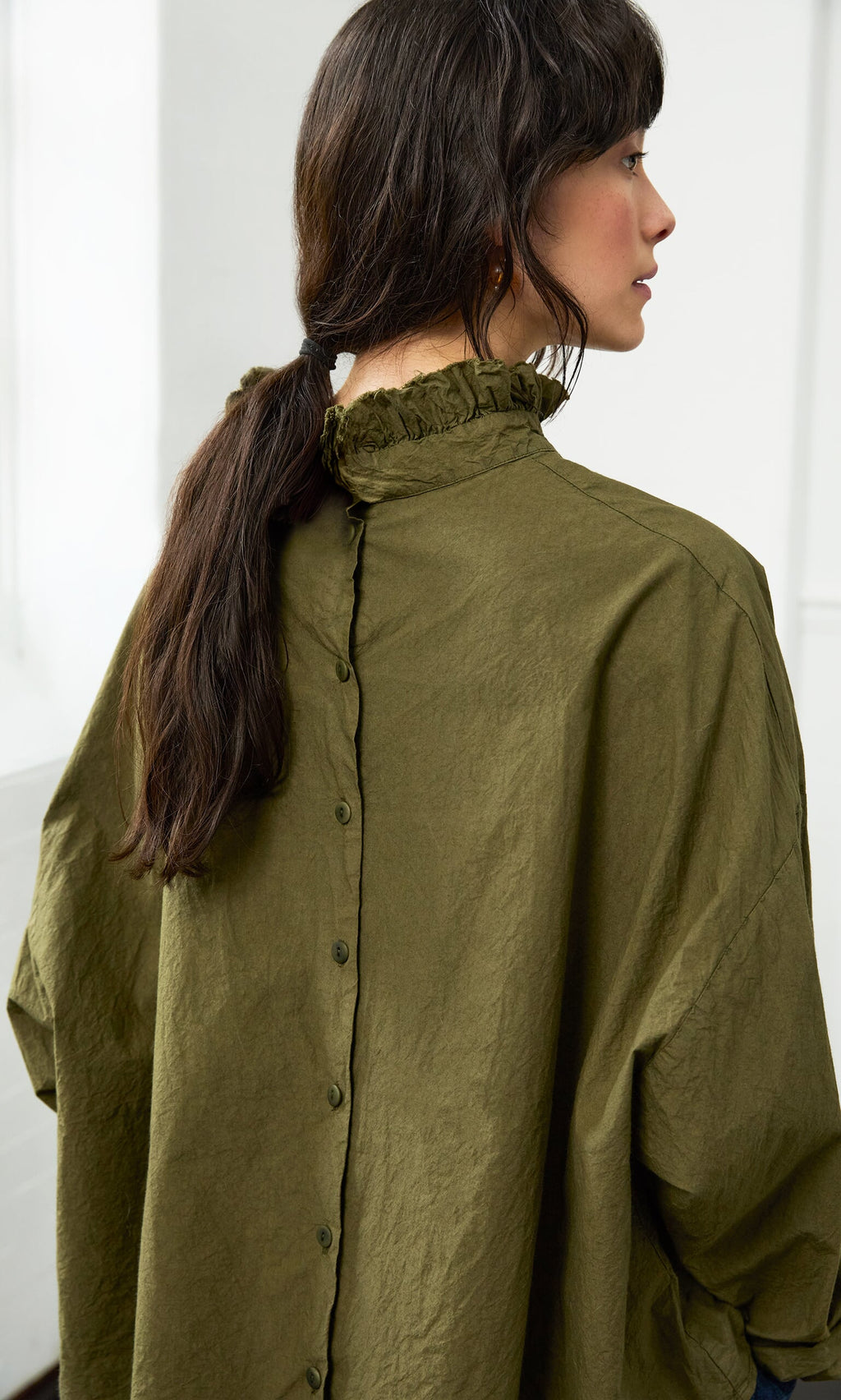 Plümo- Woman with long dark hair in the olive green Frieda Blouse featuring a ruffle neck and back buttons, seen from behind.