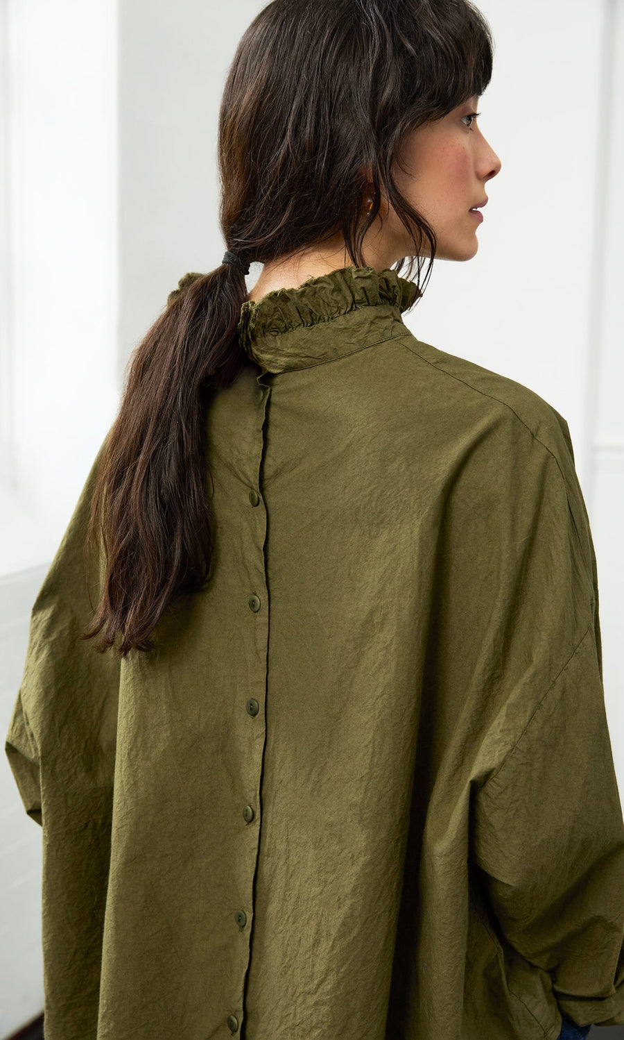 Plümo- Woman with long dark hair in the olive green Frieda Blouse featuring a ruffle neck and back buttons, seen from behind.