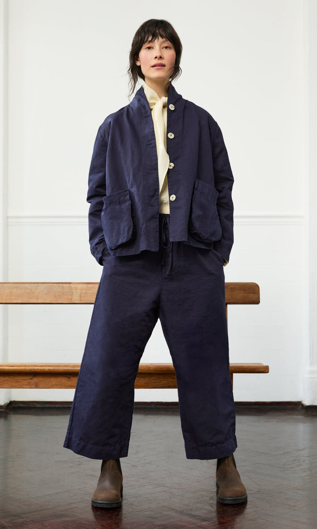 Plümo- Person indoors in a Gardener's Jacket - Navy, cotton twill wide-leg pants, cream shirt, and brown boots, hands in pockets.