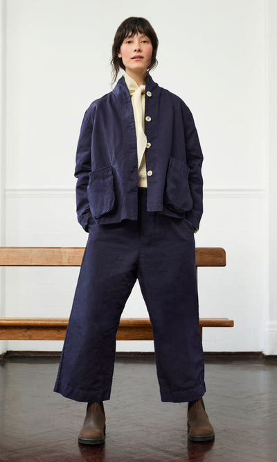 Plümo- A person stands indoors in navy Cromer Pants, a navy blue jacket, and brown boots, hands in pockets.