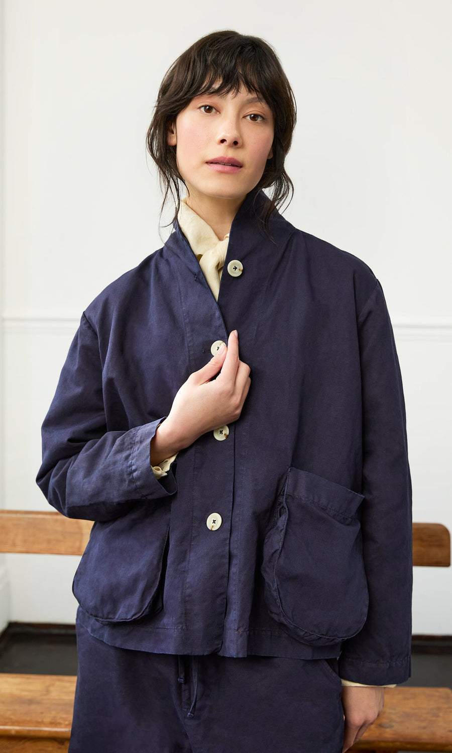 Plümo- A woman wears the Gardener's Jacket - Navy over a cream shirt, standing indoors with a neutral expression.