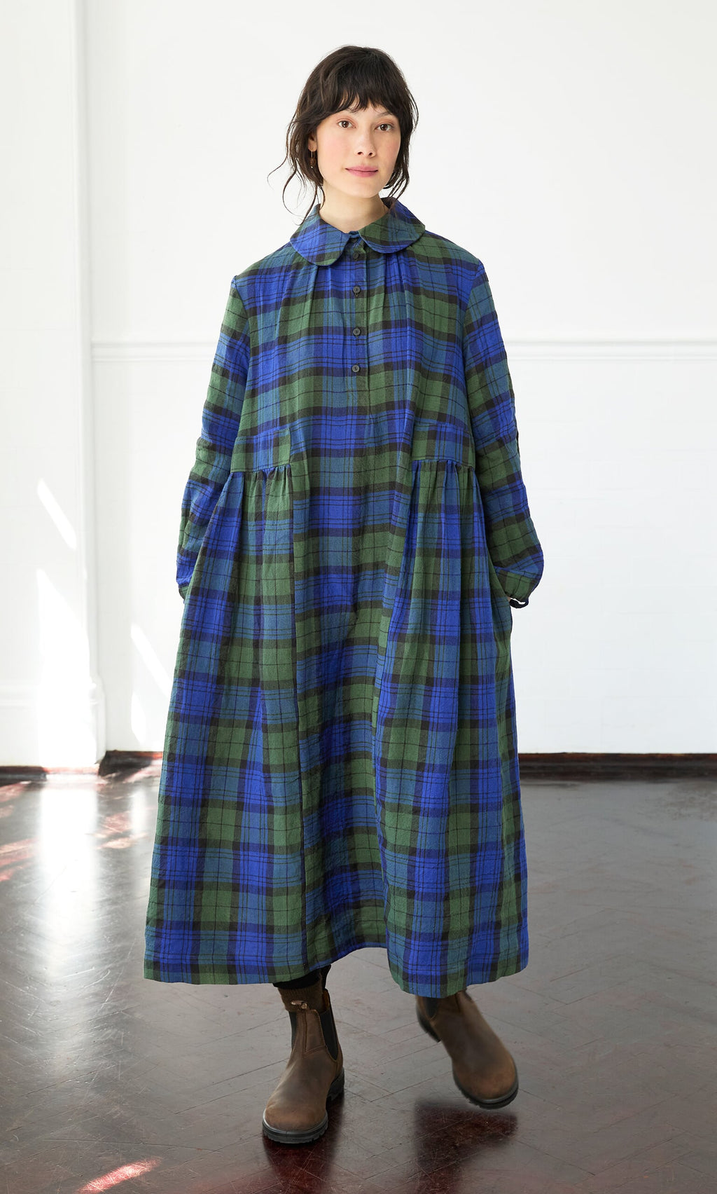Plümo- A person wears the Laois Tartan Dress—a long blue and green plaid dress with a peter pan collar—standing indoors.