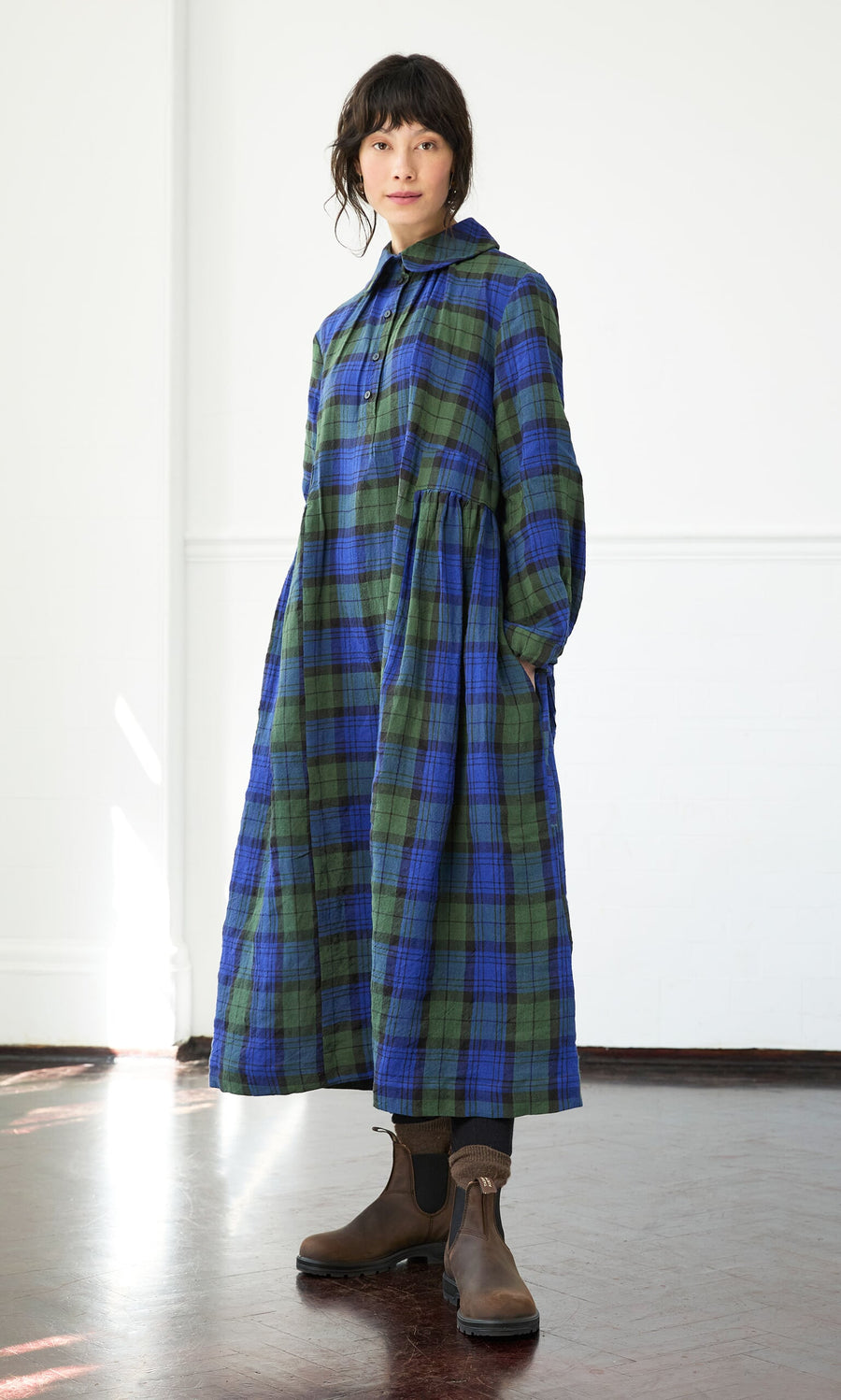 Plümo- Person wearing the Laois Tartan Dress with a peter pan collar and brown boots, standing in a bright room.