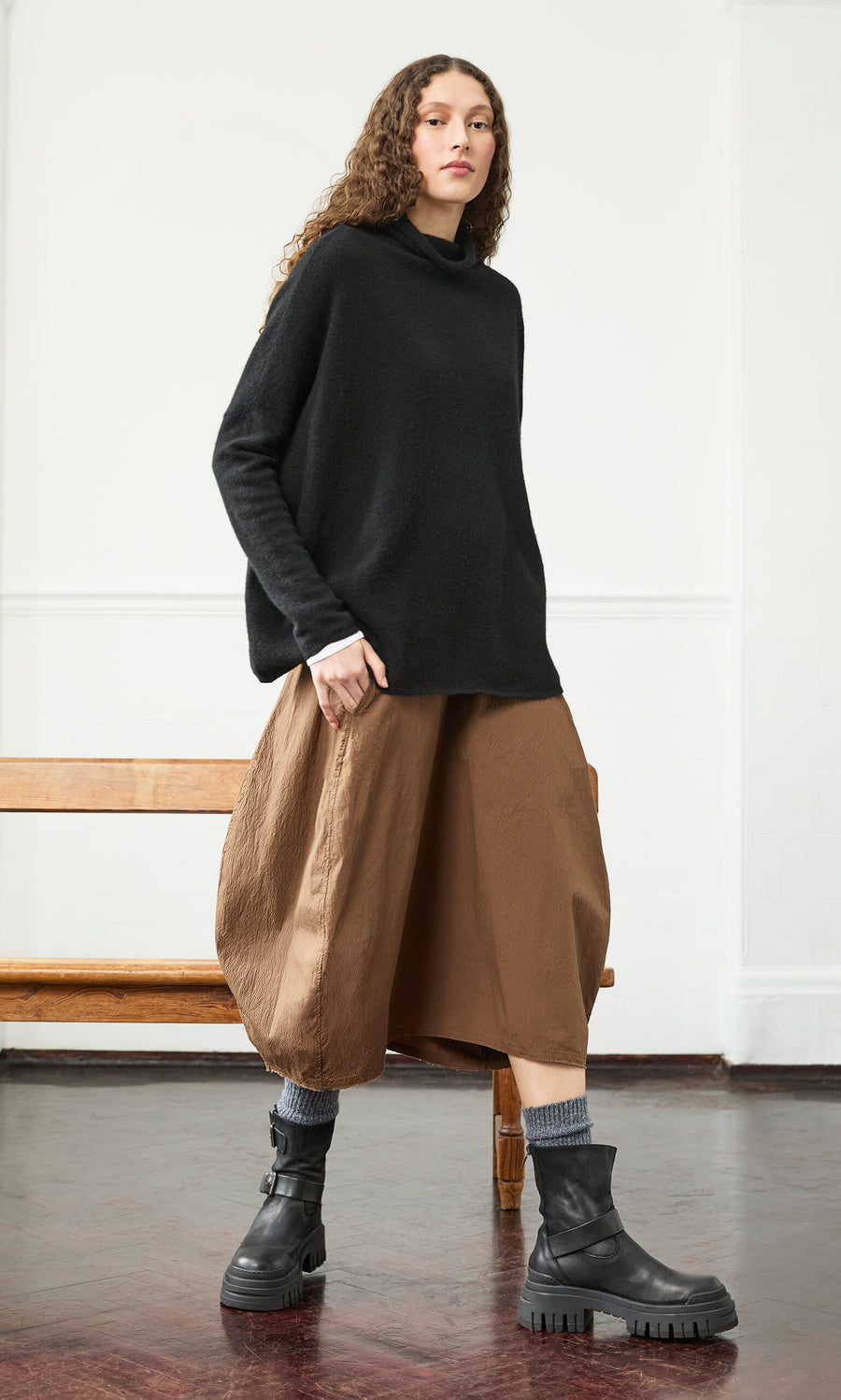 Plümo- Woman in an Onyx Rollneck, tan wide-leg pants, and black boots stands by a wooden bench in a minimalist room.