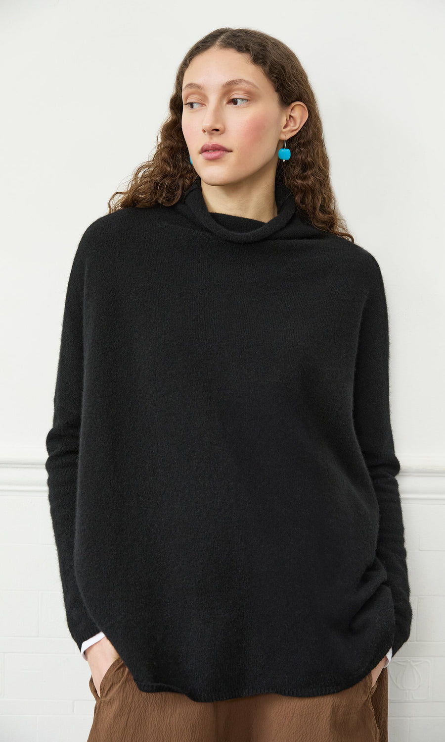 Plümo- Woman with curly brown hair wears the Onyx Rollneck, blue earrings, and brown pants against a white wall.