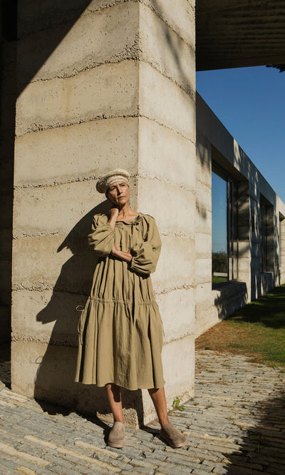 Plümo- Person in the Aiko Dress and a beret stands against a concrete wall, casting a shadow in sunlight.