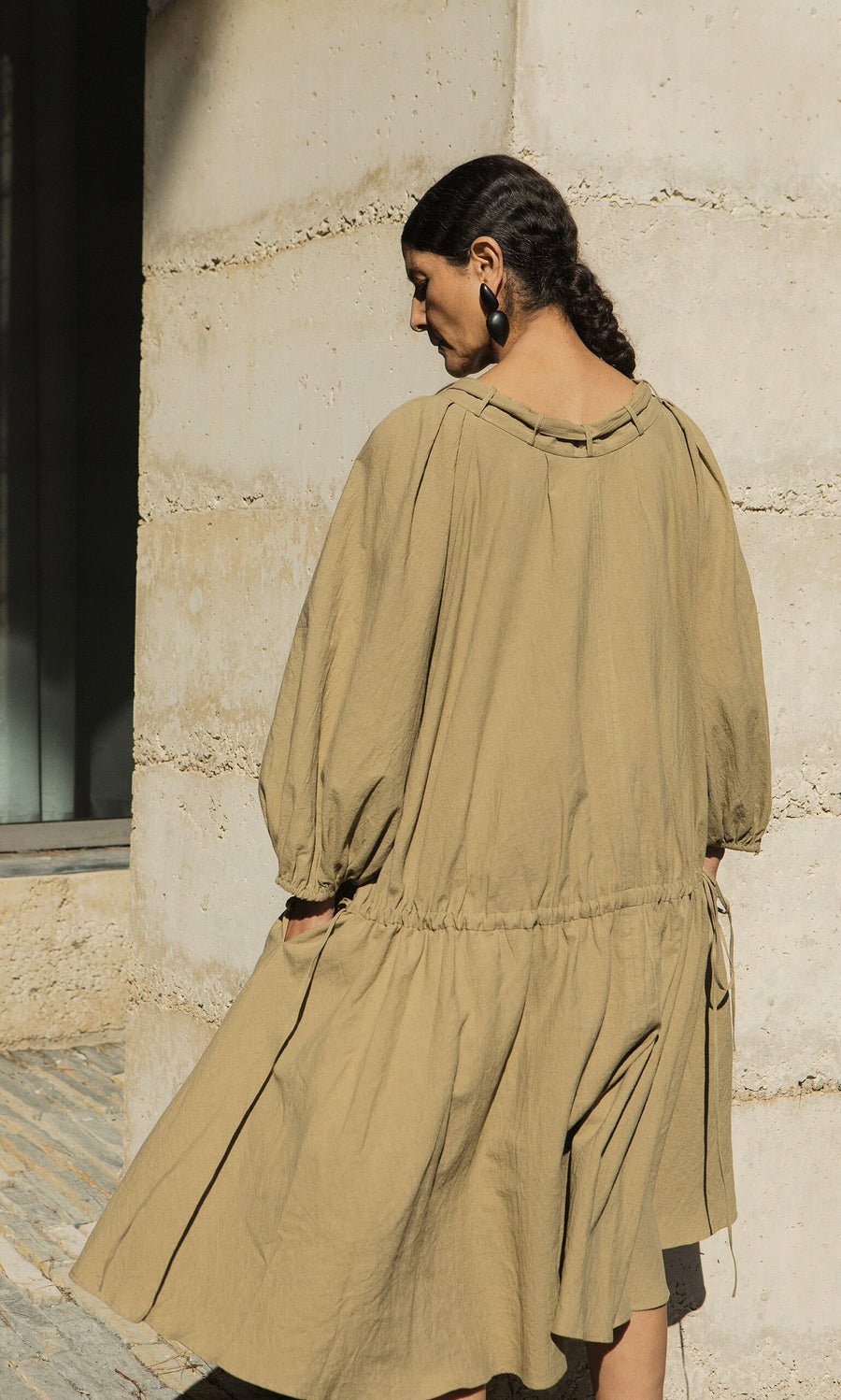 Plümo- Woman with dark hair in a low braid wears the Aiko Dress, an olive linen style with an adjustable neckline by a concrete wall.