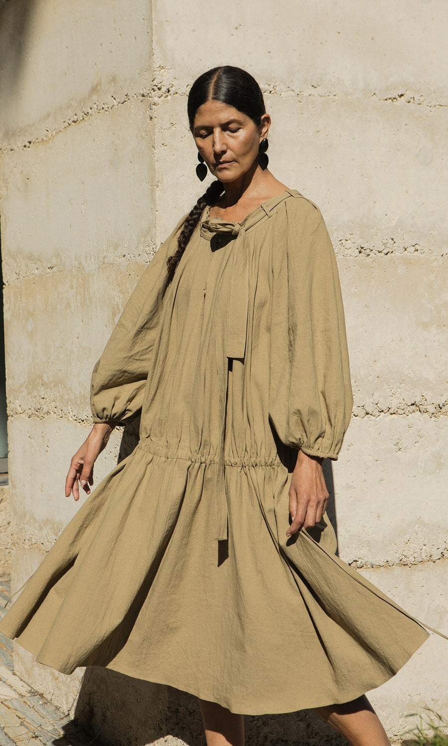 Plümo- Woman wears the Aiko Dress, a loose linen style with adjustable neckline, standing by a sunlit, textured wall.