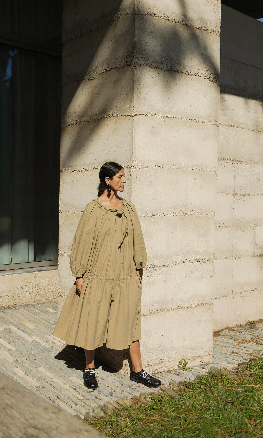 Plümo- A woman in the Aiko Dress with an adjustable neckline stands by a textured wall in sunlight, looking to the side.