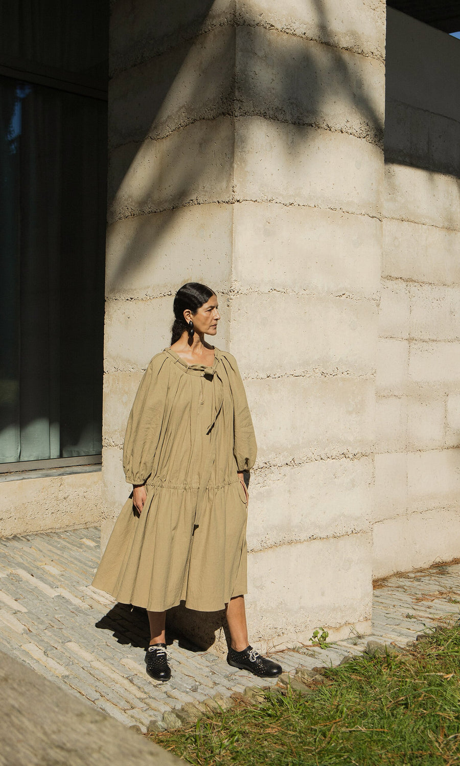 Plümo- A woman in the Aiko Dress with an adjustable neckline stands by a textured wall in sunlight, looking to the side.