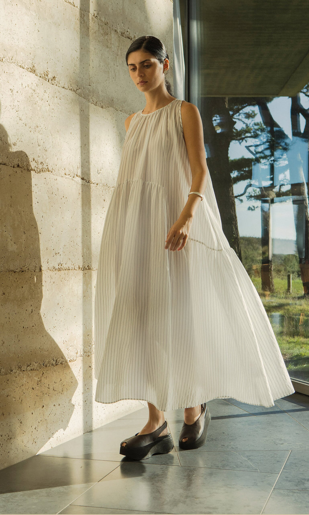 Plümo- Woman wearing the Alba Stripe Dress stands indoors by a sunlit textured wall and glass window.