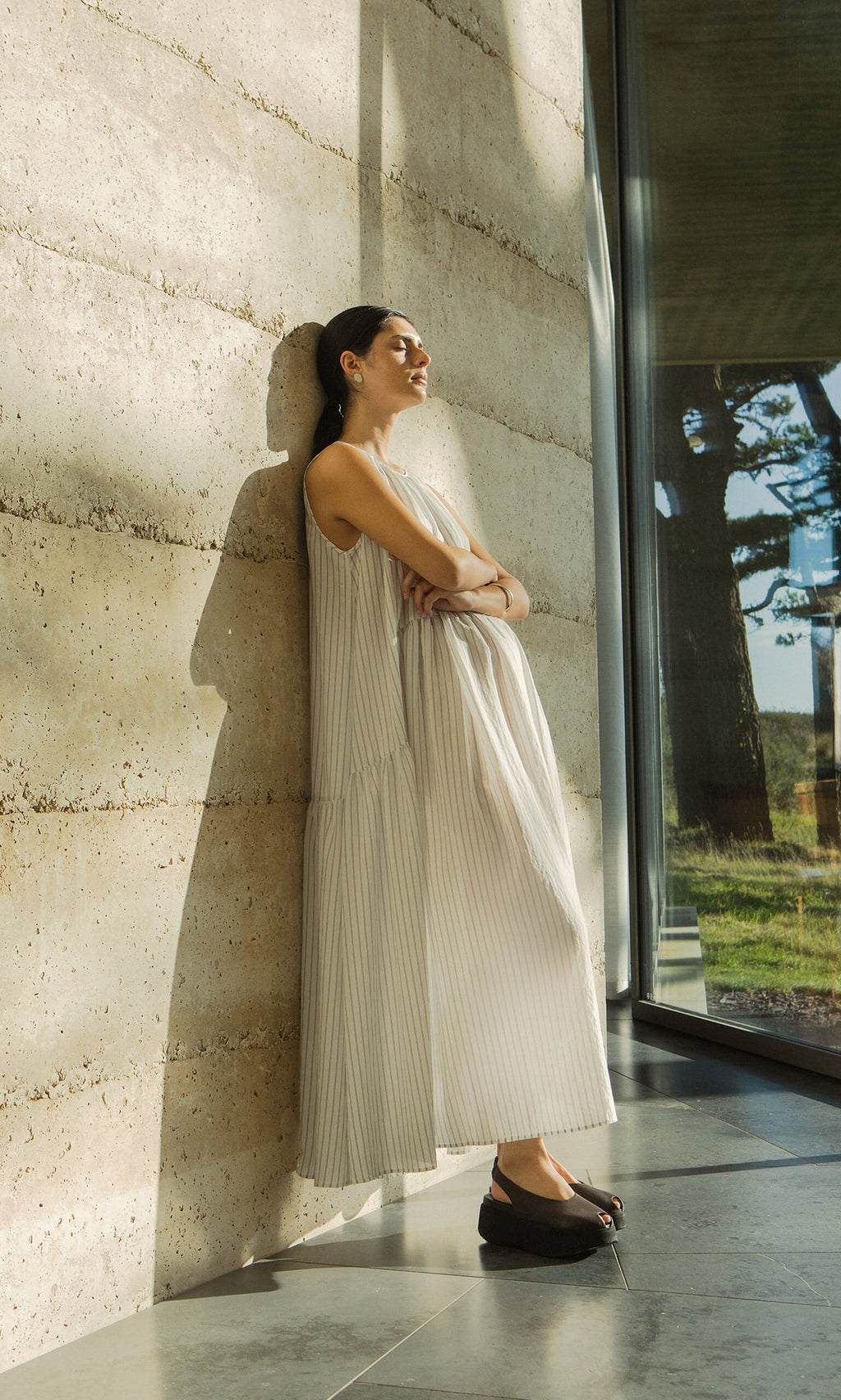 Plümo- Woman in the sleeveless Alba Stripe Dress leans against a sunlit wall by a window, eyes closed and arms crossed.