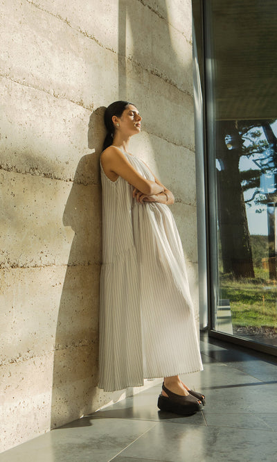 Plümo- Woman in the sleeveless Alba Stripe Dress leans against a sunlit wall by a window, eyes closed and arms crossed.