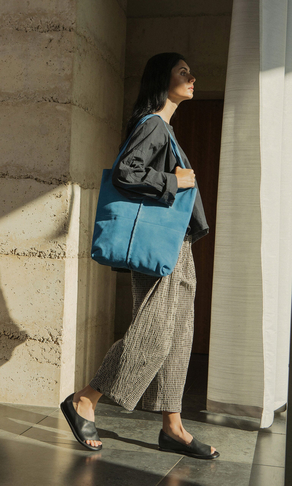 Plümo- Woman in patterned pants and black jacket walks indoors, carrying a blue tote and wearing Hawke Sandals.