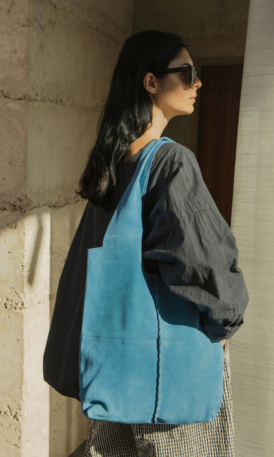 Plümo- Woman with long dark hair in sunglasses, loose dark top, carrying the Alves Bag—a blue suede shoulder bag.