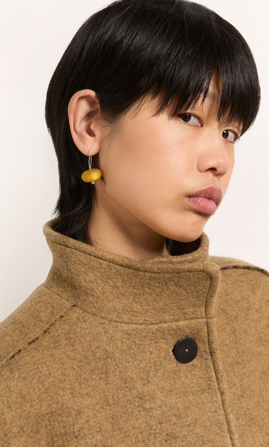 Plümo  Person with straight black hair wears a tan coat and Bola Earrings - Honey Dew, looking sideways at the camera.