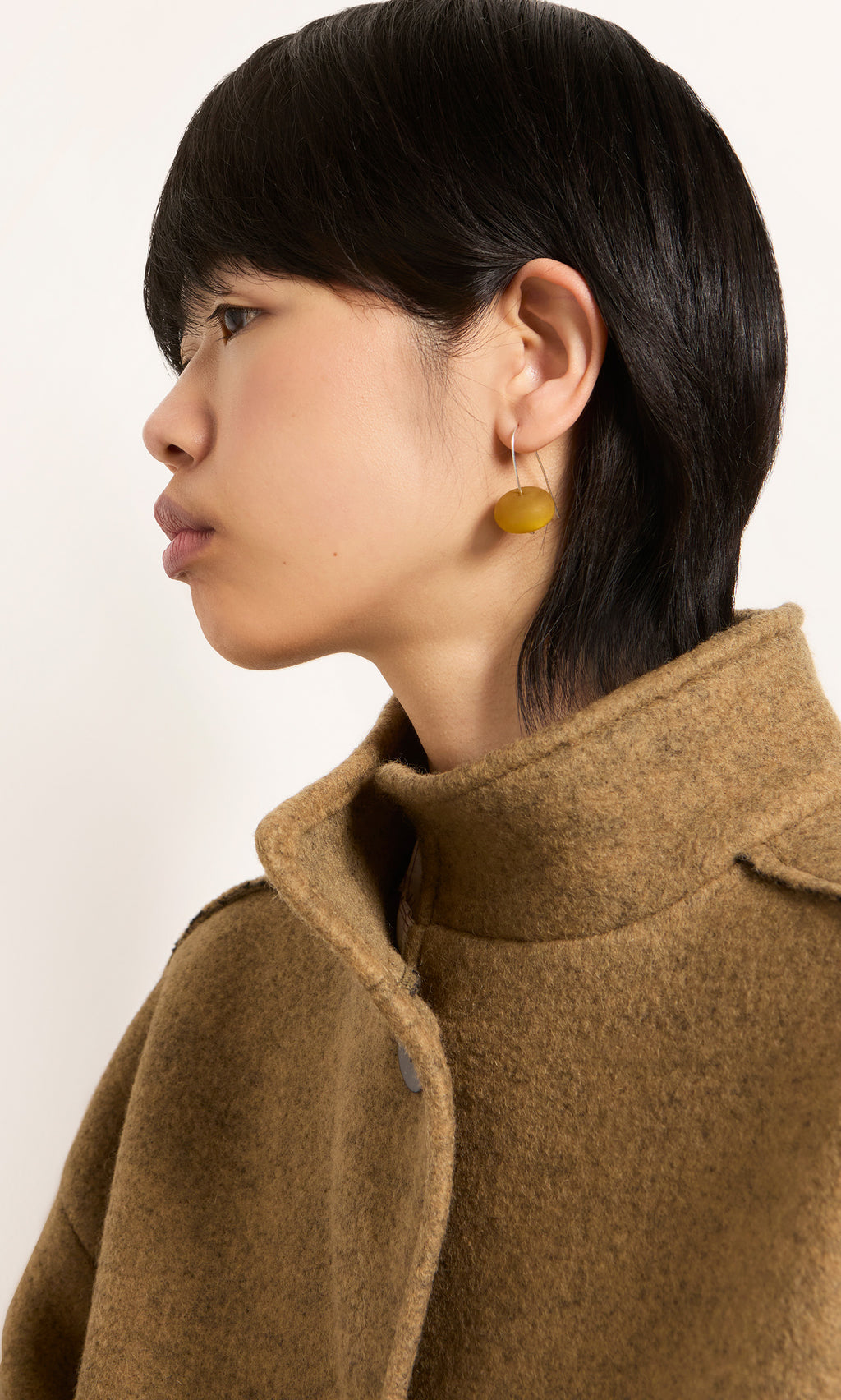 Plümo  A person in a brown coat looks left, showing Bola Earrings - Honey Dew with short dark hair and a neutral look.