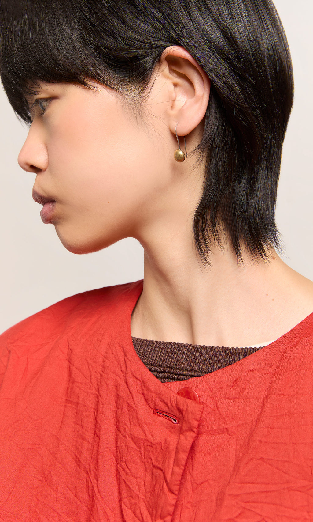 Plümo  Person in red top with short dark hair, facing left, wearing Greta Earrings - Brass small gold recycled brass earrings.