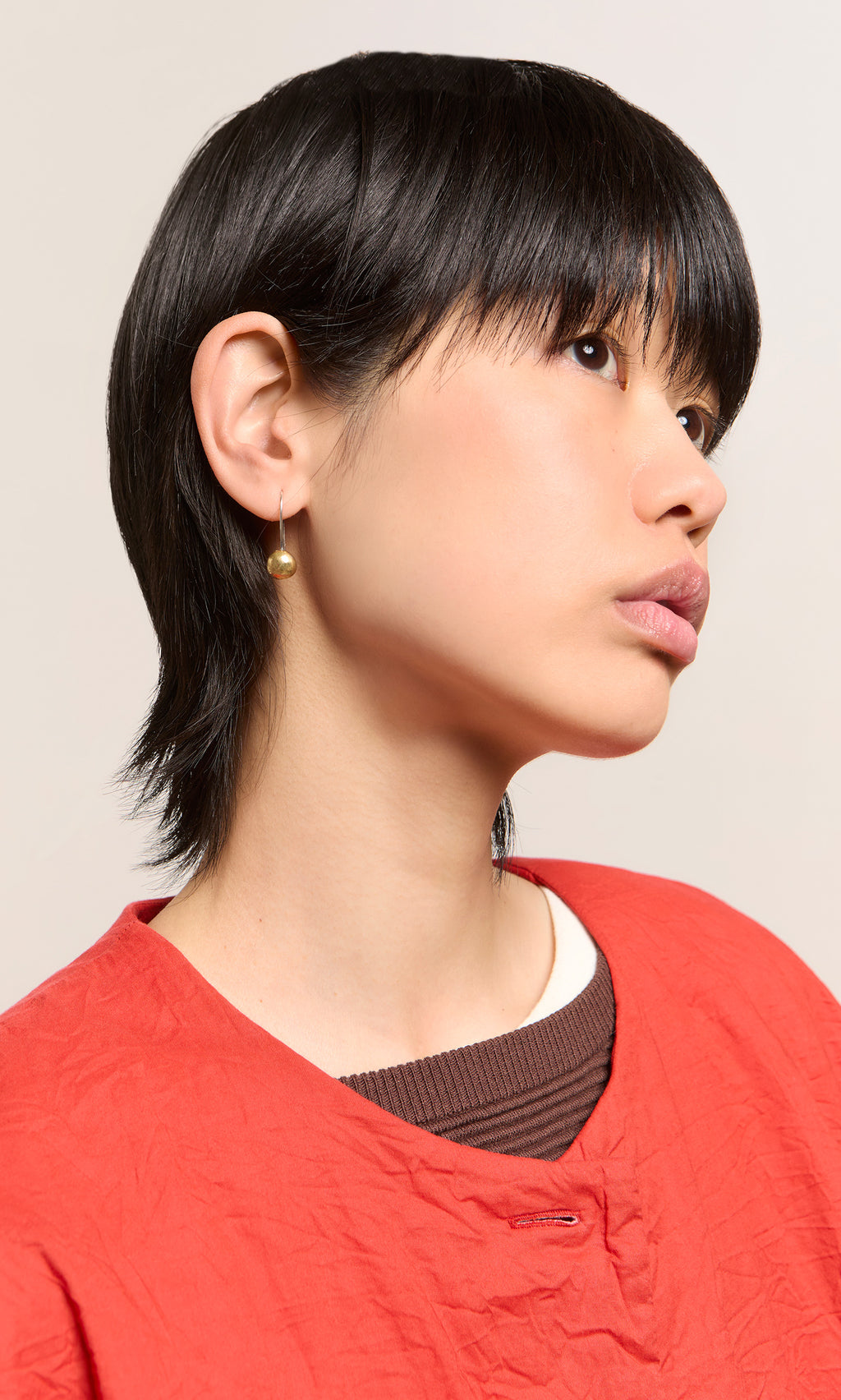 Plümo  Person with short black hair in a red top wears Greta Earrings - Brass, looking upward against a plain background.