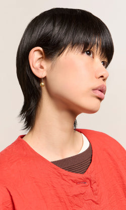 Plümo  Person with short black hair in a red top wears Greta Earrings - Brass, looking upward against a plain background.