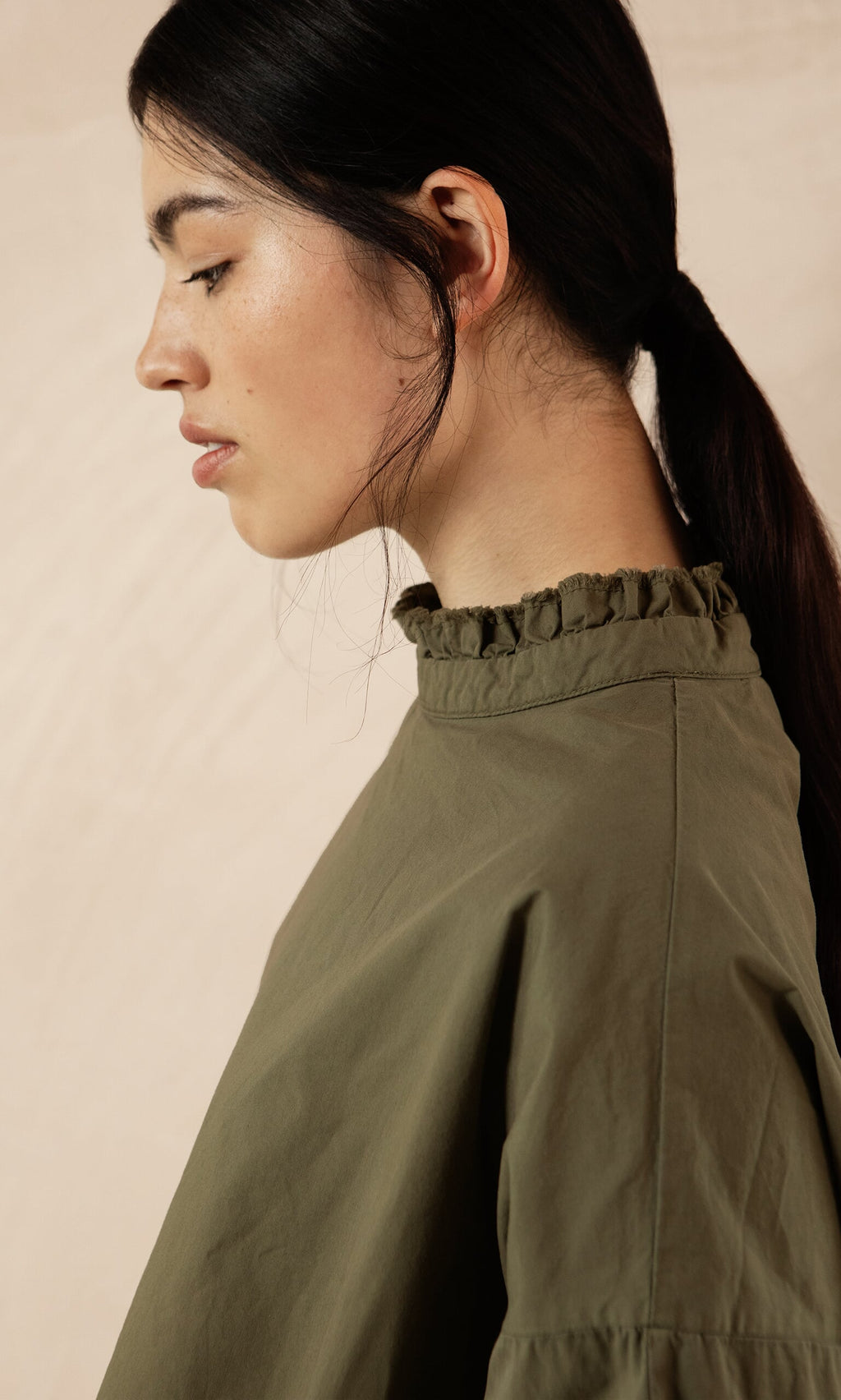Plümo- Woman with dark hair in a low ponytail wears the Olive Ruffle Blouse, shown in side profile.