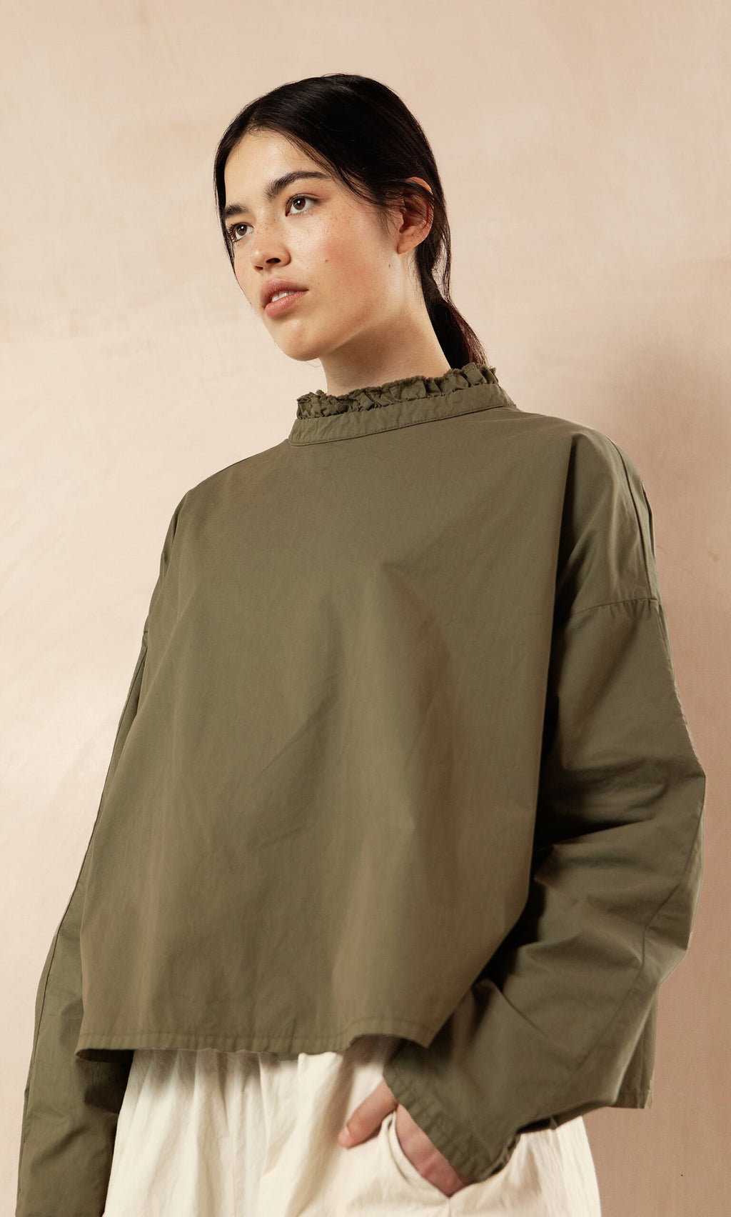 Plümo- Woman wearing an Olive Ruffle Blouse and white pants, standing against a beige background.