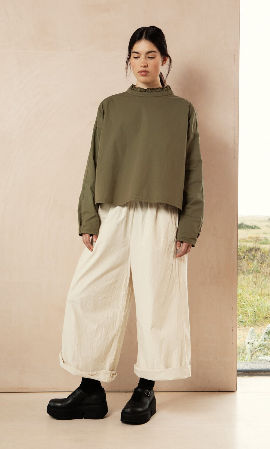 Plümo- Woman in an Olive Ruffle Blouse, white wide-leg pants, and black shoes stands against a beige wall.