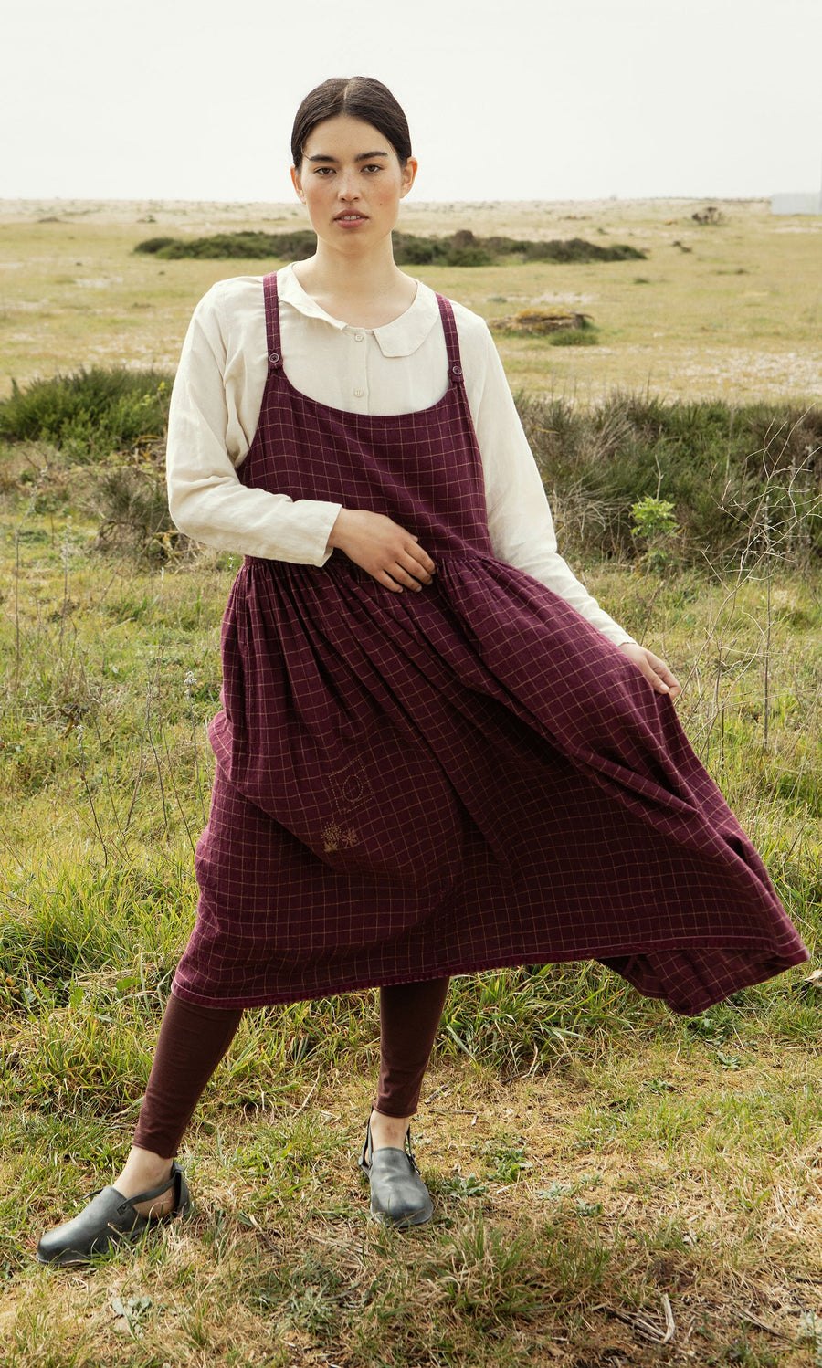 Plümo- Woman in the Anja Dress, a Swedish-designed linen apron dress with checks, stands outdoors on grass, looking at camera.
