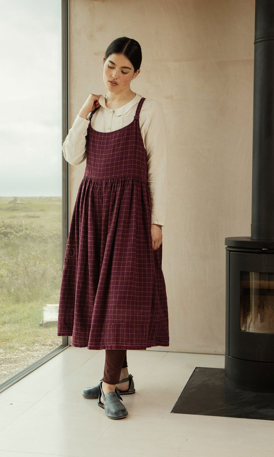 Plümo- Woman in the Anja Dress, a burgundy chequered pinafore, over a white blouse stands by a window and black fireplace.