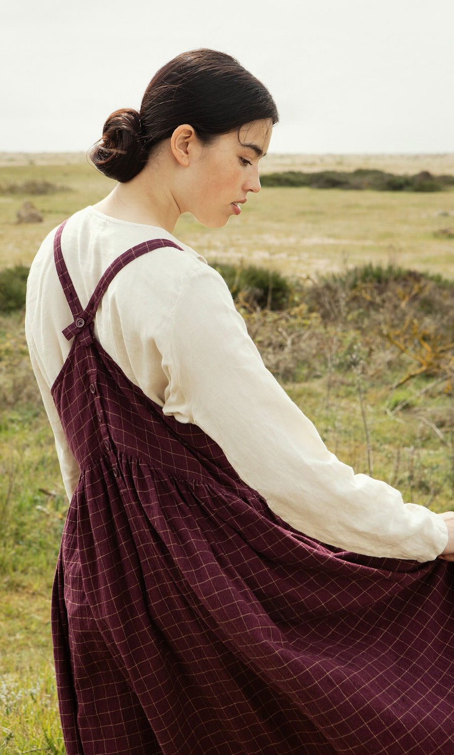Plümo- A woman in a cream shirt and the Anja Dress stands in a grassy field, looking downward.