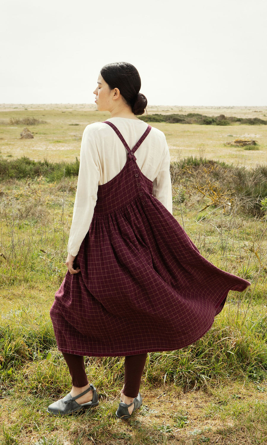 Plümo- Woman wearing the Anja Dress, a maroon linen apron dress with chequered pattern, standing in a windy grassy field.