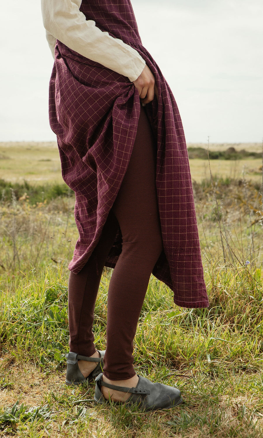 Plümo- A person in historical clothing lifts an Anja Dress, revealing brown leggings and black shoes outdoors.